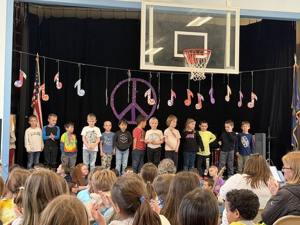 A row of young students stands on stage during a school concert while an audience of students and adults watches and applauds.