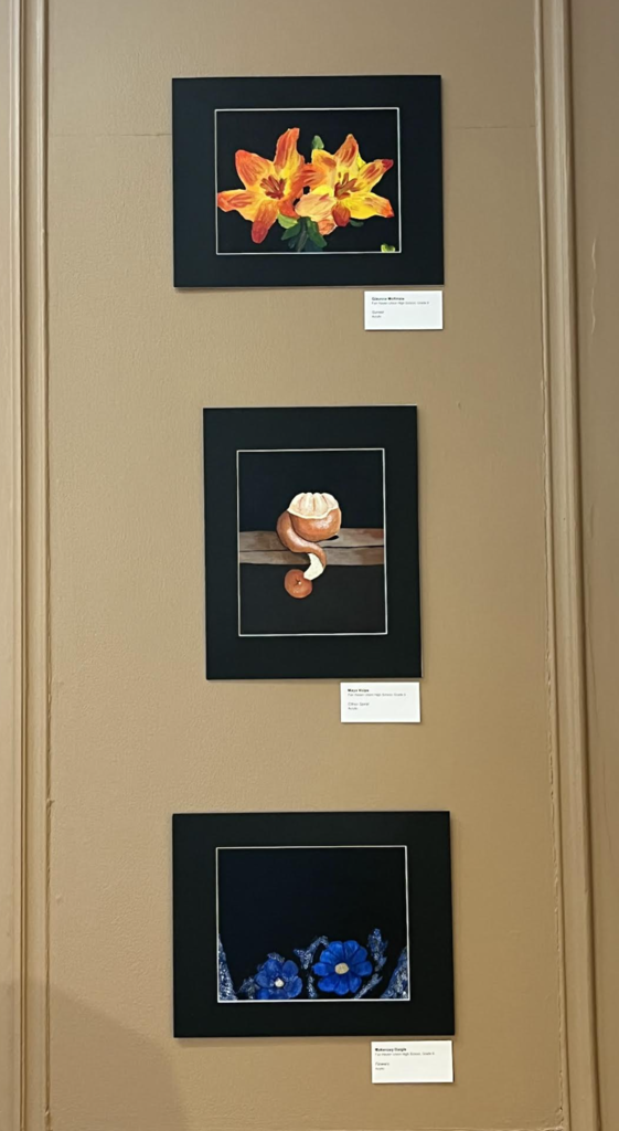 Three framed student artworks on a wall, including a painting of orange flowers, a still life of peeled fruit, and blue flowers on a dark background.