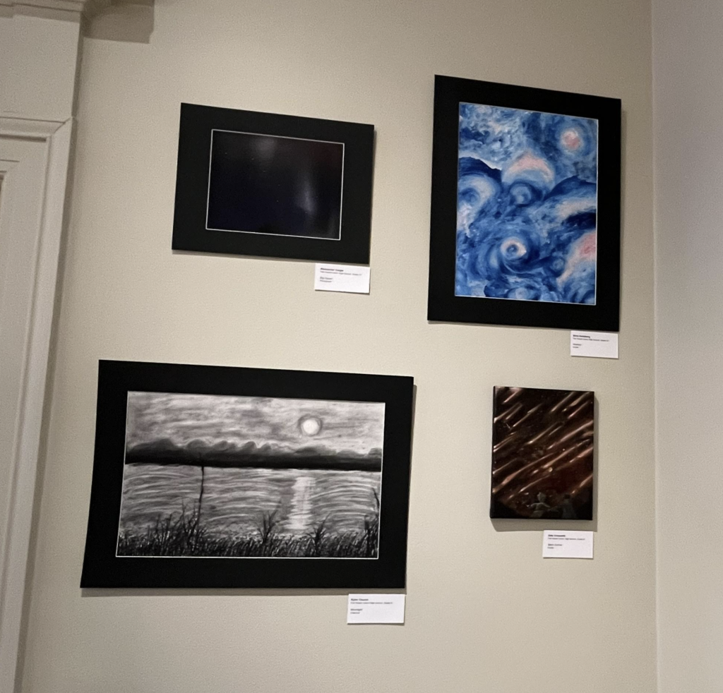 Gallery wall featuring several student artworks, including a blue abstract painting, a black-and-white landscape drawing, and a small textured piece.