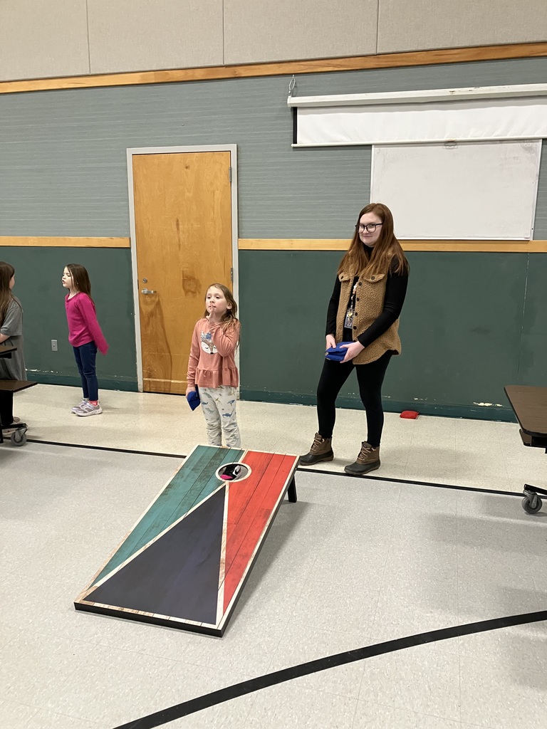  A young girl stands ready to play a game of cornhole in a gymnasium or community room. She holds a blue beanbag and looks toward the camera. A woman in a brown fleece vest stands behind her, and another child is visible in the background. The cornhole board features a geometric design with teal, red, and black sections.  