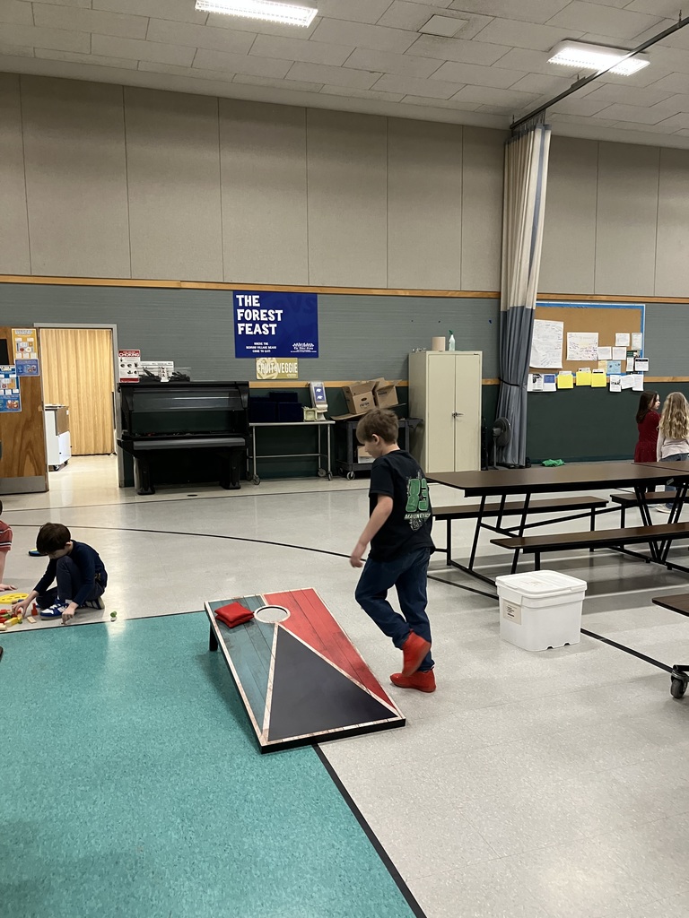 A wide shot of a multi-purpose room where several children are playing. In the foreground, a boy walks past a cornhole board toward the center of the room. To the left, another child sits on the floor playing with wooden blocks. A "The Forest Feast" poster hangs on the wall near a piano and various storage cabinets.  