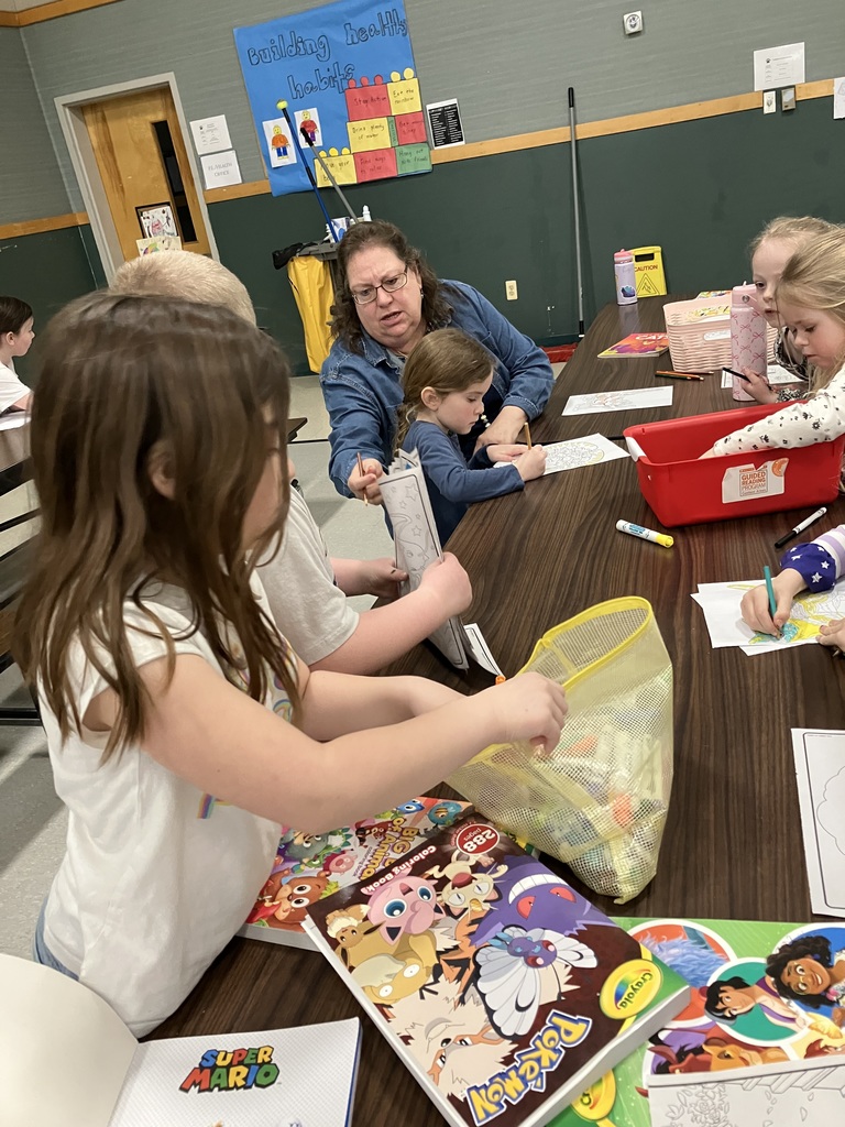  An indoor scene showing a group of young children and an adult seated at a long wooden table. The children are focused on coloring books, including Pokémon and Super Mario themes. A woman in a denim shirt leans over to assist a young girl with her coloring. In the background, a colorful "Building Healthy Habits" poster is visible on the wall.  