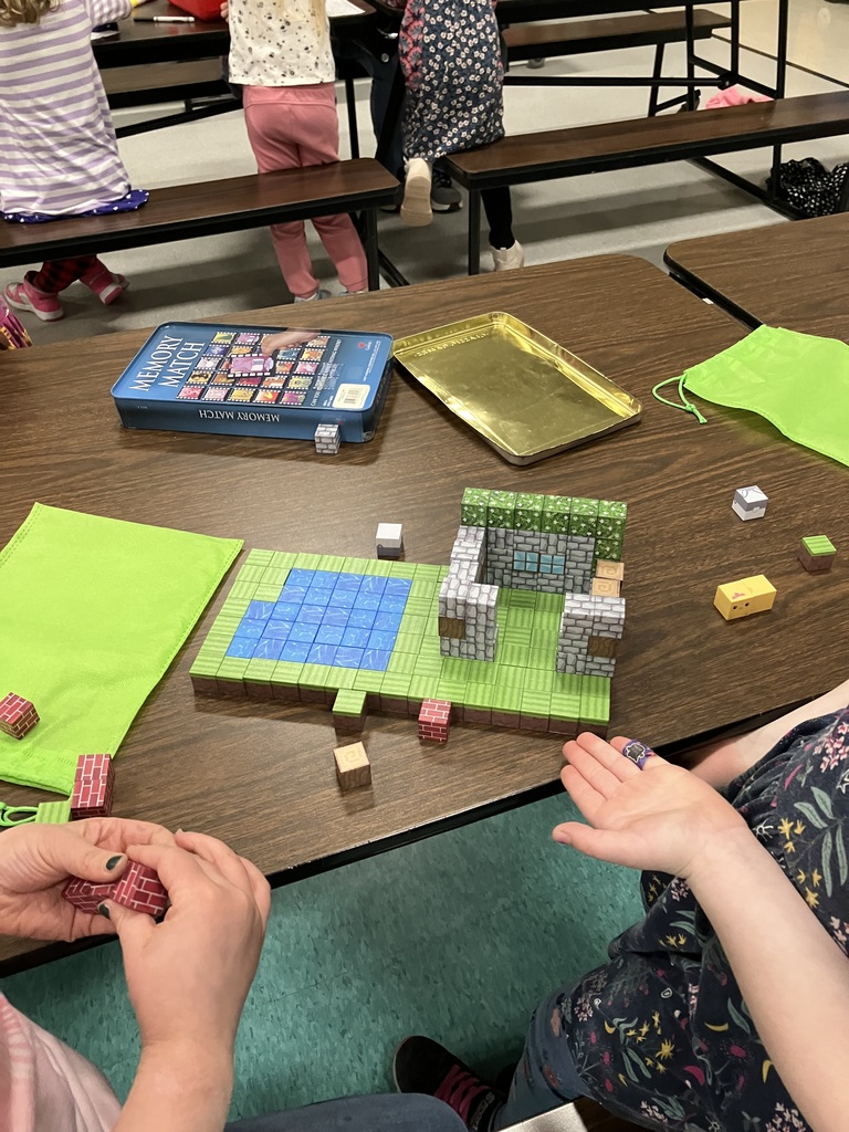 A close-up, high-angle shot of a wooden table where children are playing with magnetic building blocks that resemble Minecraft textures. A small structure with stone-patterned walls and a green roof stands on a base of grass and water tiles. Nearby, there is a blue "Memory Match" game tin and a gold metal tray.  