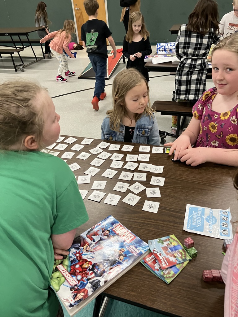 Three young girls sit at a table engaged in different activities. Two girls in the foreground are looking at a large array of face-down memory match cards spread across the table. Next to them is a Marvel-themed activity book and more magnetic building blocks. Other children and a cornhole game are visible in the blurred background.