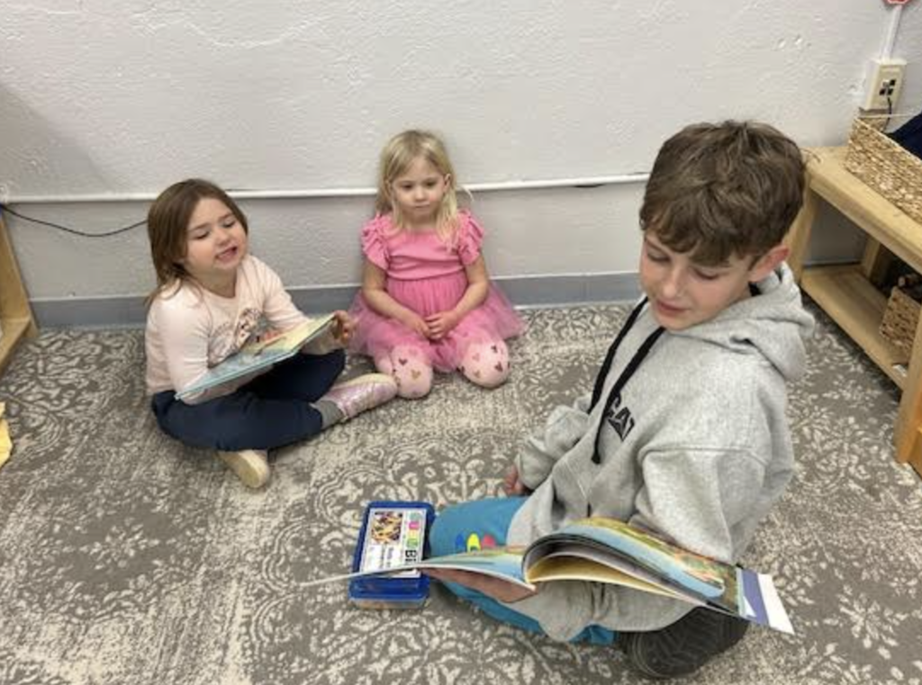 Three students sit on the floor reading books together near a classroom shelf and learning materials.