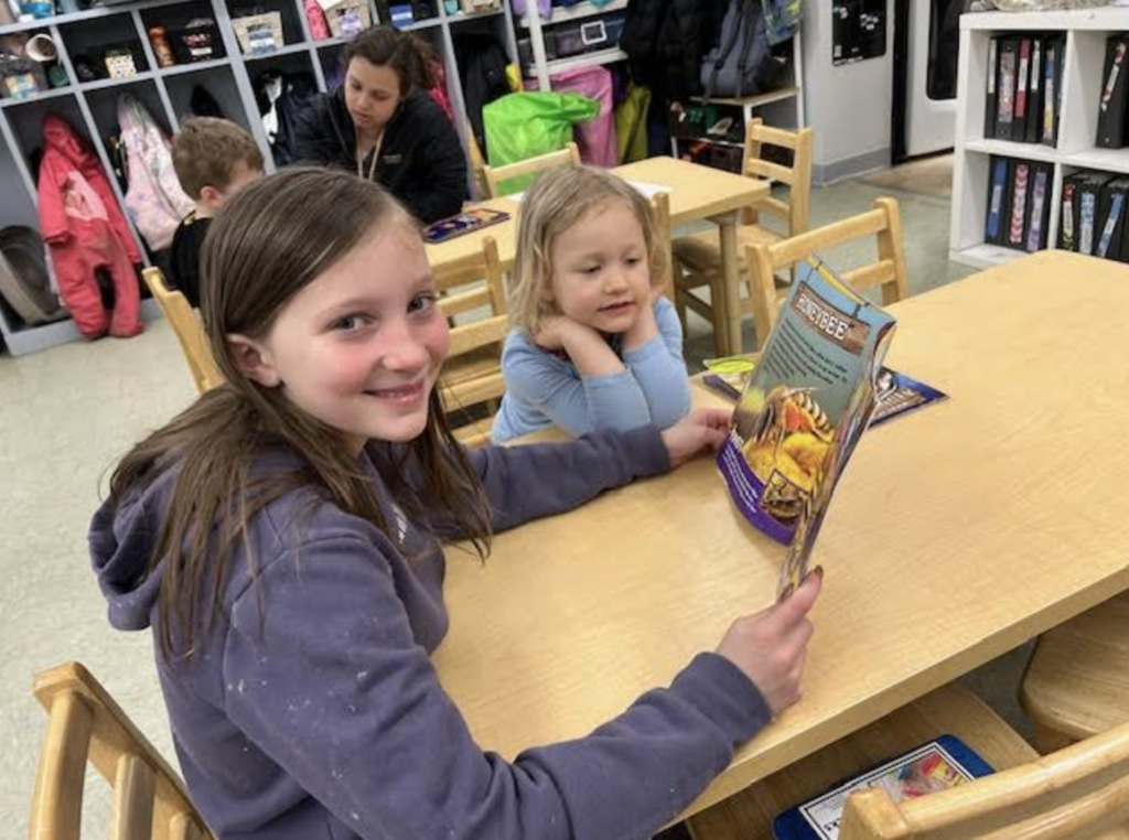 Two students sit at a small table reading a book together while an adult assists children in the background.