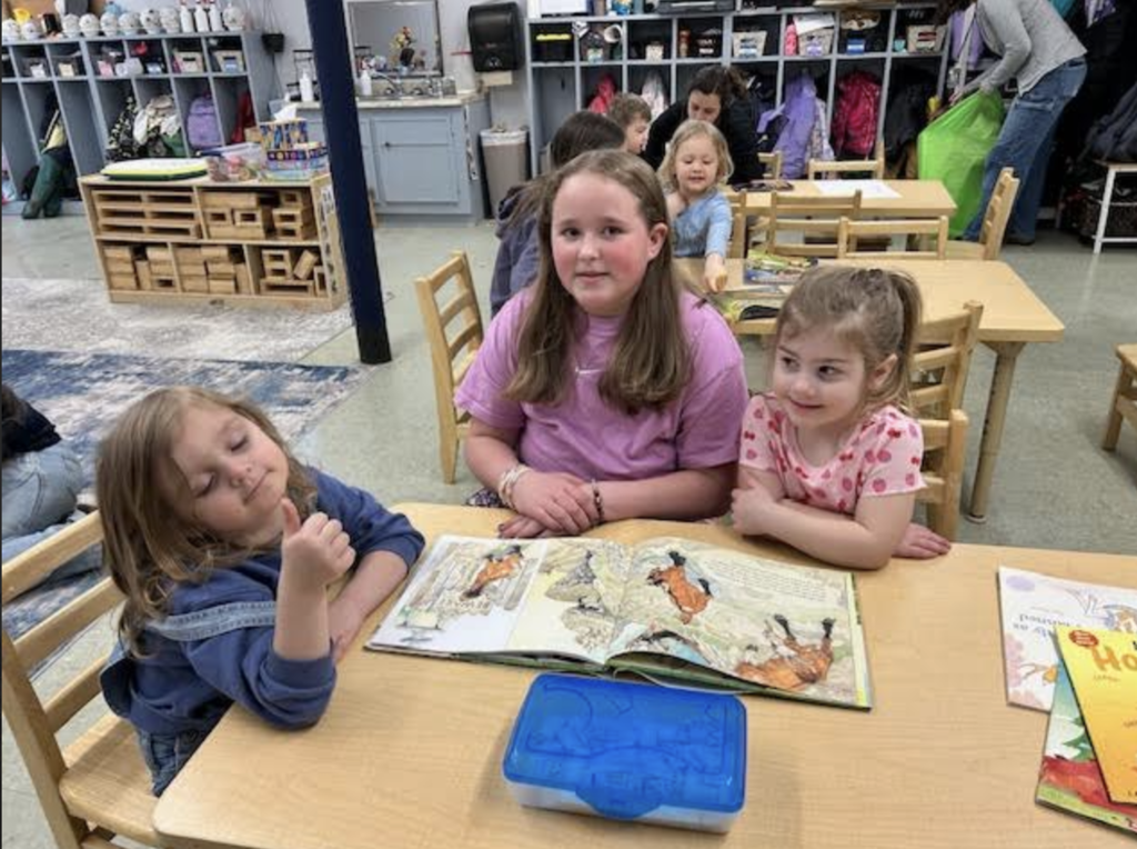 Three students sit at a table looking at an open picture book while other children work in the classroom behind them.