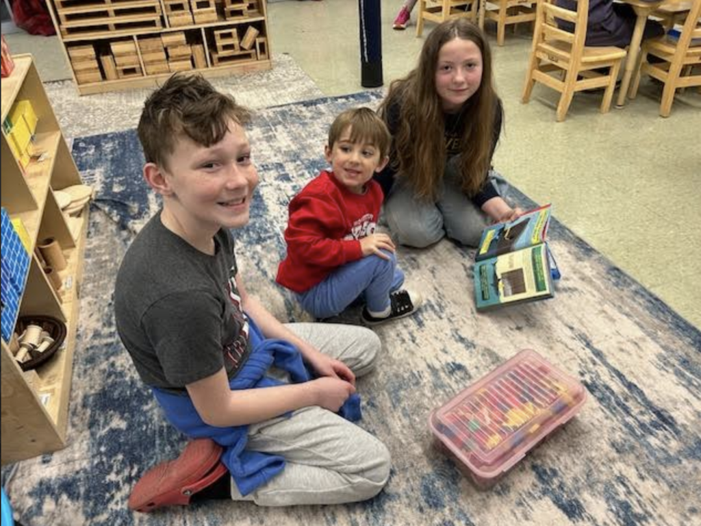 Three students sit on a classroom rug smiling and reading together with books and art supplies nearby.