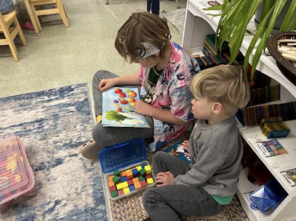 Two students sit on the floor reading a picture book together next to a container of colorful blocks.