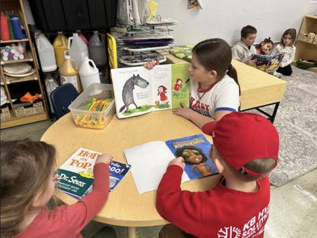 A student reads a picture book aloud to two classmates seated at a round table with other books and classroom supplies.