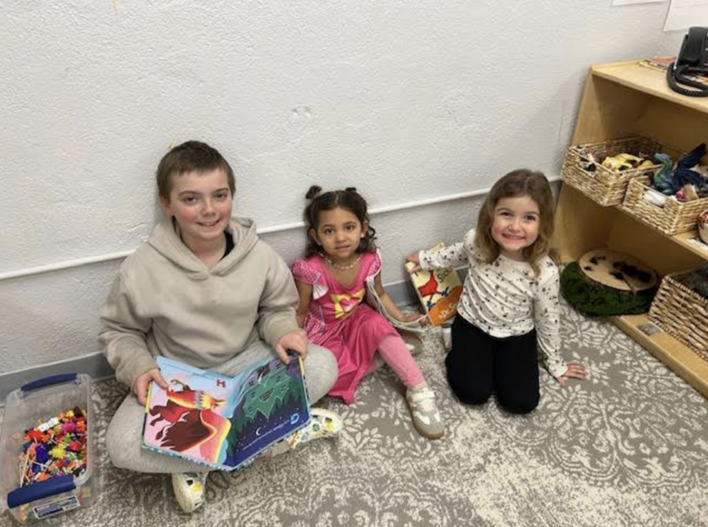 Three young students sit on the floor reading books together beside a classroom shelf with toys and materials.