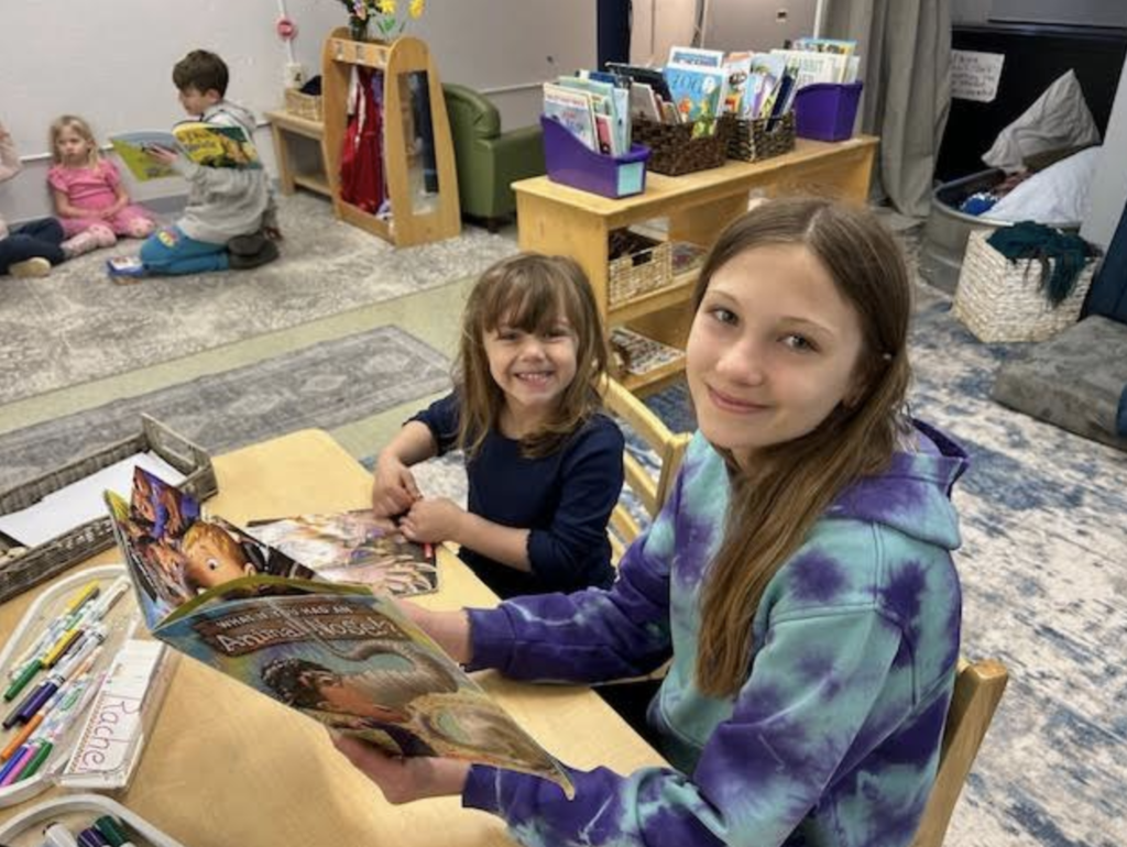 Two students sit at a table smiling while reading books, with a classroom library and other students visible in the background.