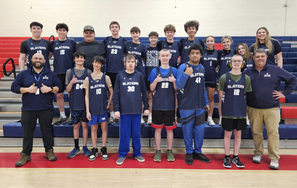 Group of high school Unified basketball players and coaches pose in a gym wearing Slaters jerseys, standing in front of bleachers and smiling together.
