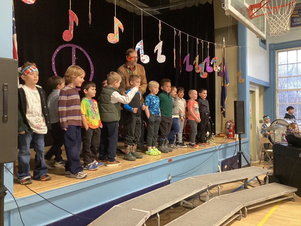 Elementary students stand on a stage with a teacher, decorated with colorful music notes and a peace sign for a school performance.