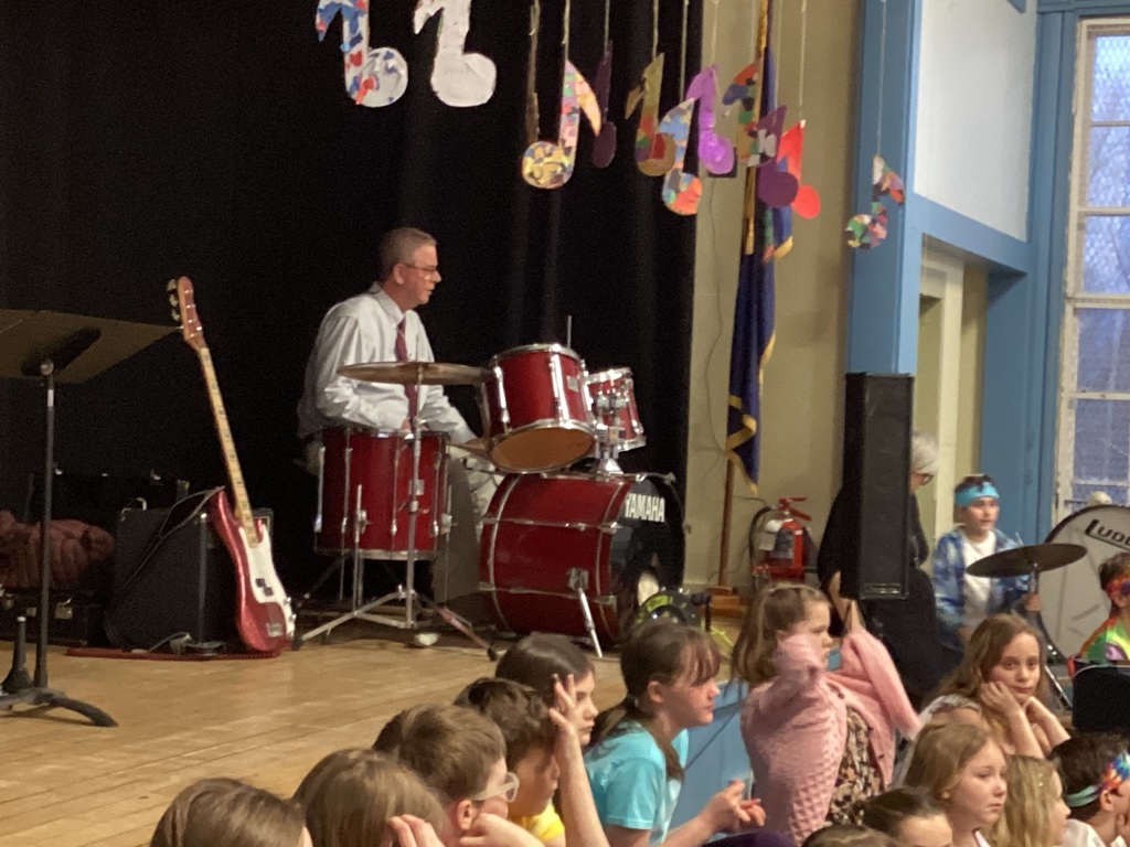 Adult drummer plays a drum set on stage while students sit nearby during a school concert.