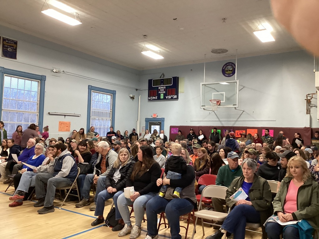 Crowd of families and community members seated in a school gymnasium, facing forward for a student performance.