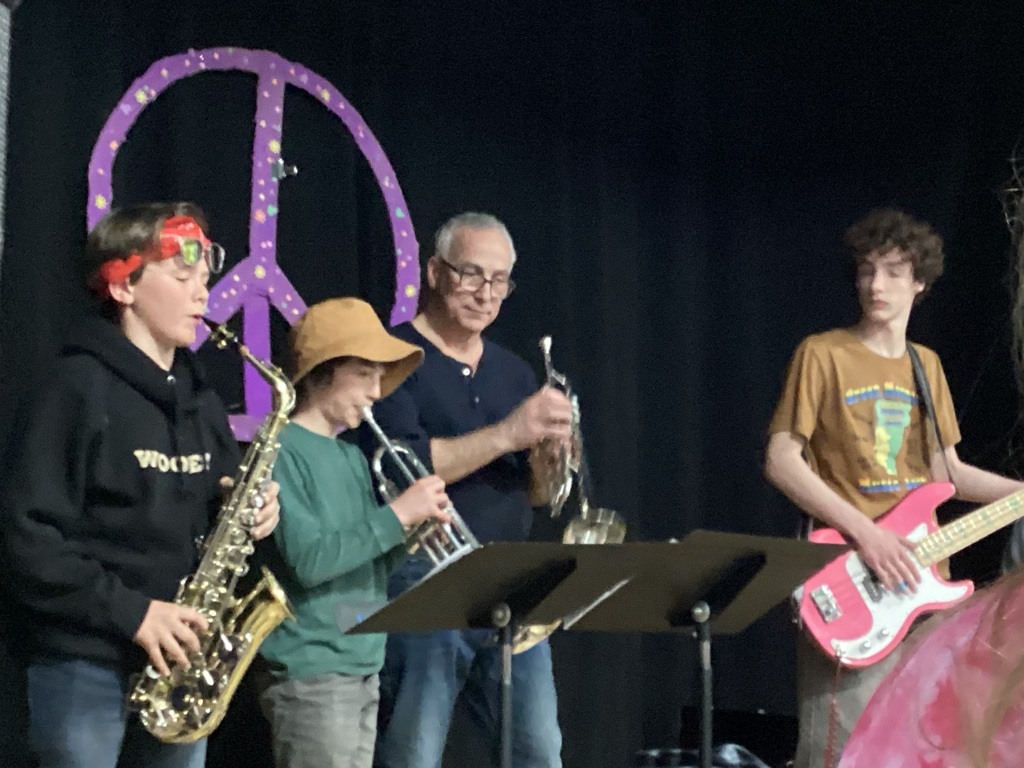 Small group of students and an adult play saxophone, trumpet, and bass guitar on stage during a live performance.