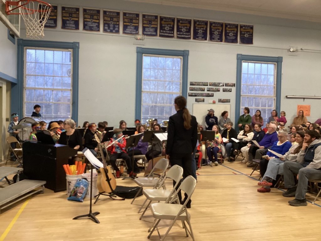 School band performs in a gymnasium as a conductor leads student musicians, with audience seated nearby.