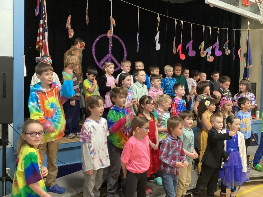 Large group of elementary students in colorful outfits singing together on stage during a school concert.
