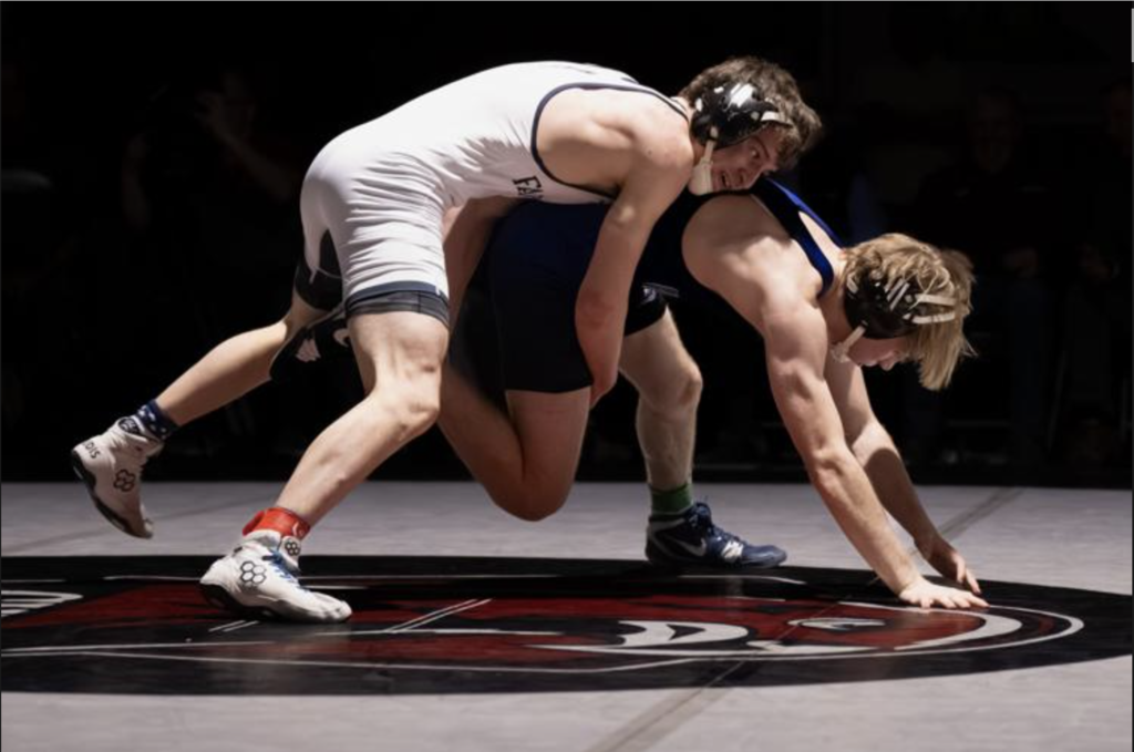 Two high school wrestlers compete on a mat, with one athlete attempting a takedown while the other braces on hands and knees.