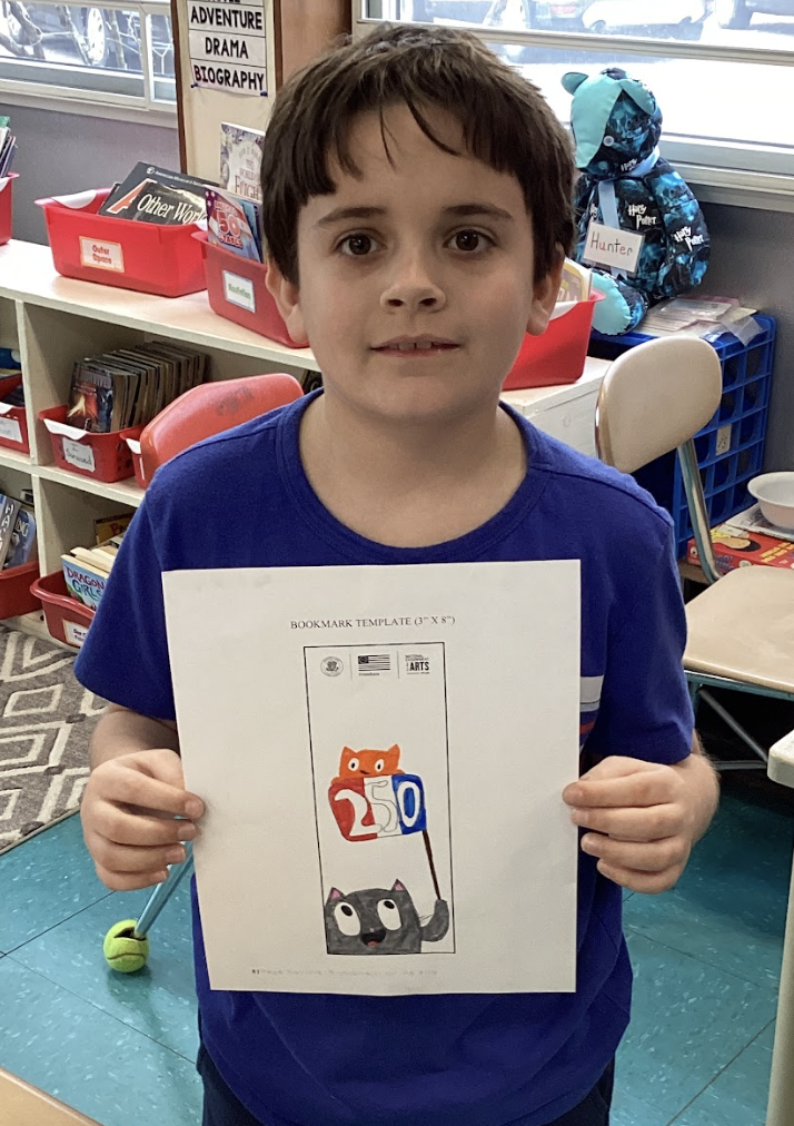 Student holds a bookmark featuring cartoon cats and the number “250,” standing in a classroom library area.