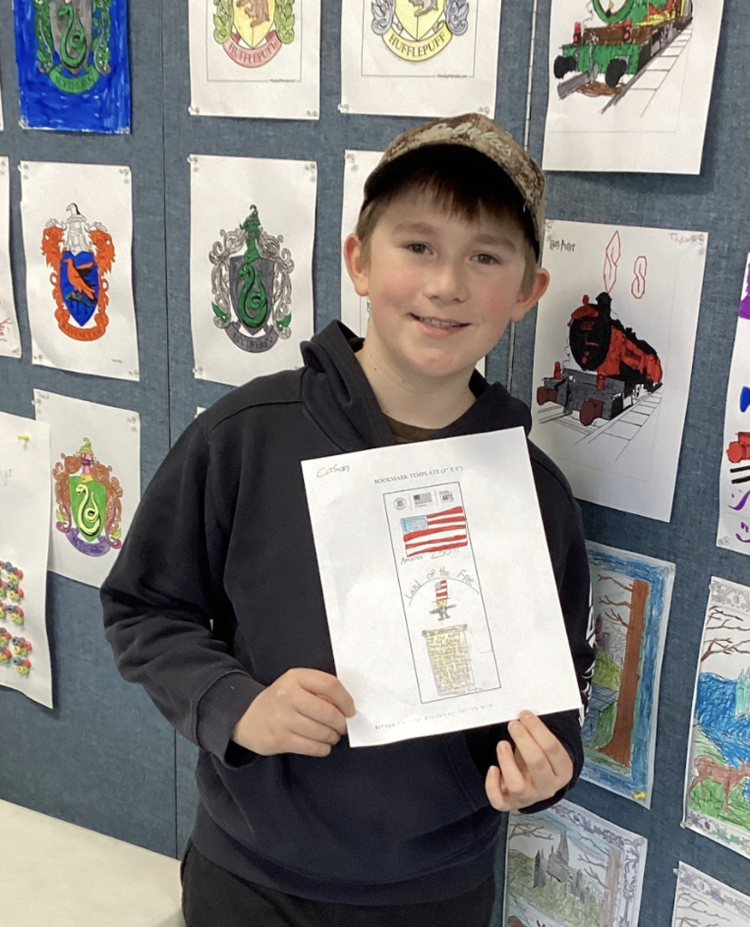 Student smiles while holding a bookmark design featuring the American flag, standing in front of a wall display of student artwork.