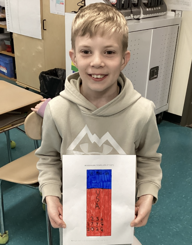 Student holds a red and blue bookmark design reading “USA 250 1776–2026” while standing in a classroom.