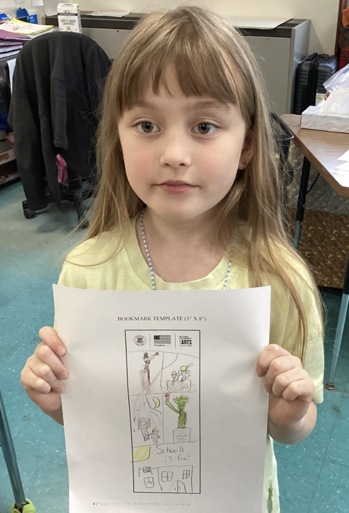 Young student holds a hand-drawn bookmark featuring multiple scenes and the words “School is fun.”