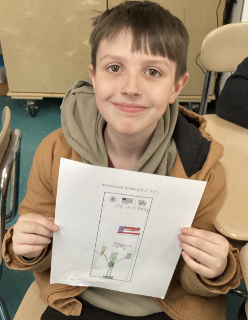 Student holds a bookmark design reading “250 years of the USA” with a flag and simple drawings.