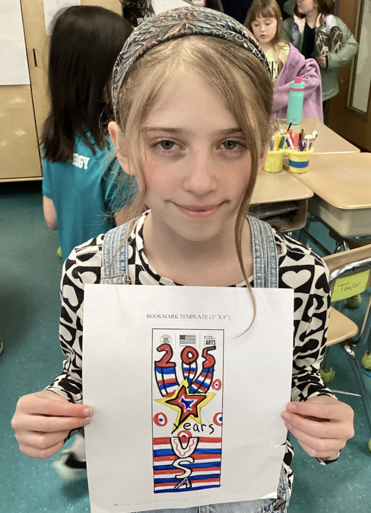 Student holds a colorful bookmark design reading “205 years USA” with stars and stripes in a classroom setting.