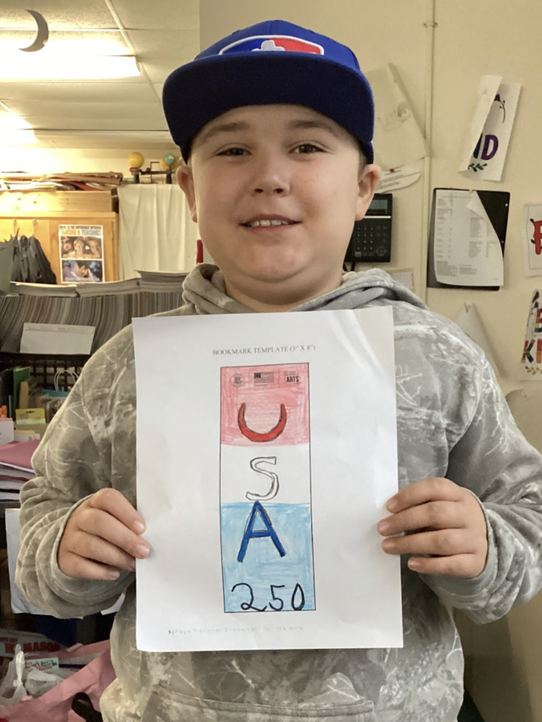 Student wearing a blue cap holds a red, white, and blue bookmark design labeled “USA 250” in a classroom.
