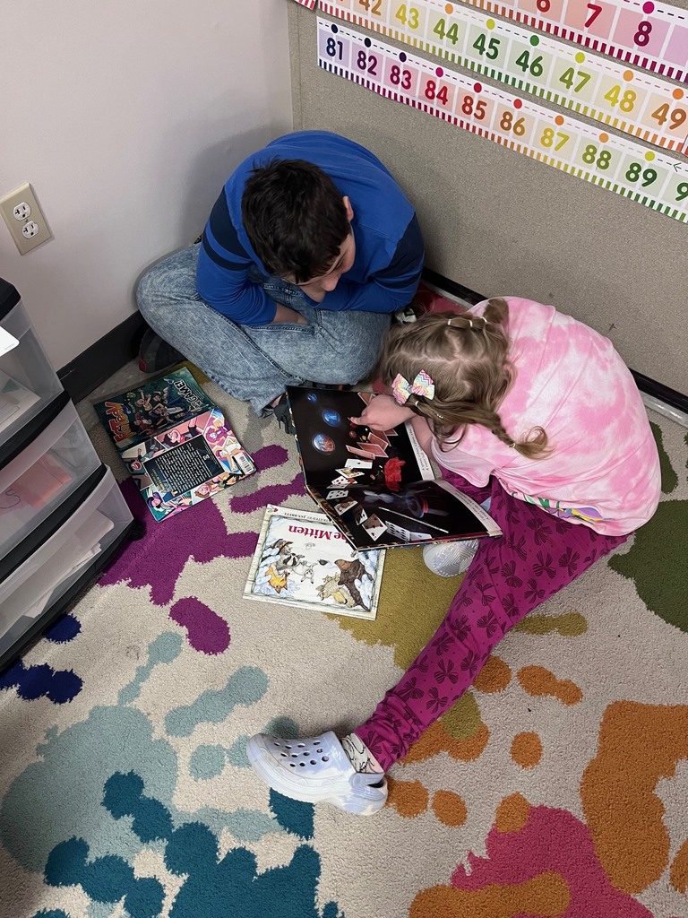 From a high angle, two children are seen sitting on a colorful, paint-splatter patterned rug. They are focused on an open picture book about magic tricks, with other books like The Mitten lying nearby.