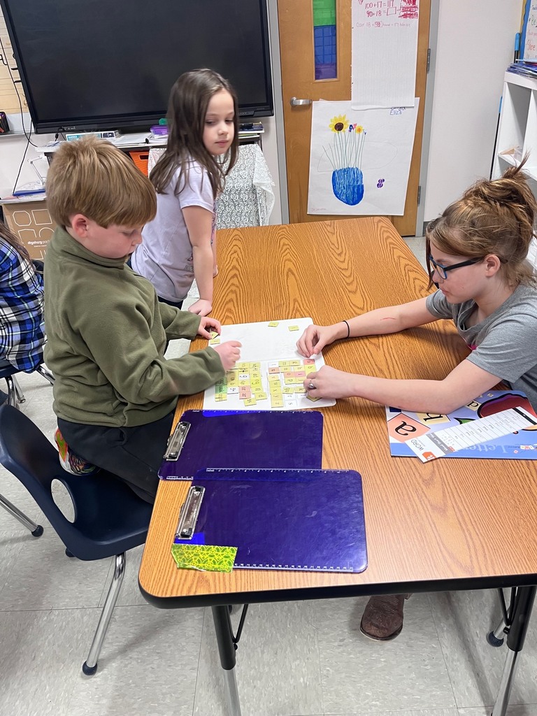 Two children sit at a wooden table working together on a word-building activity with small yellow letter tiles on a white board. A third child stands nearby, watching the process.