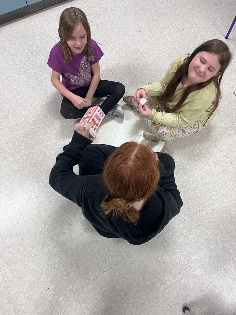: A high-angle shot of three girls sitting in a circle on the floor. One girl holds a "Pop for Sight Words" game box while another holds a small circular word tile.