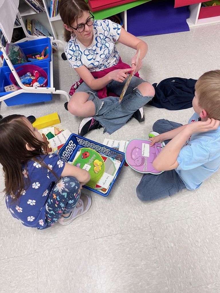 An older student sits cross-legged on the floor, guiding two younger children through a literacy game involving animal-shaped cards and word fragments in a blue plastic basket.