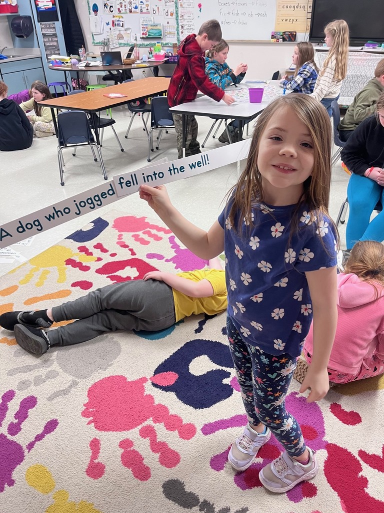 A young girl in a floral shirt smiles at the camera while holding up a long white sentence strip that reads, "A dog who jogged fell into the well!" Other children are active in the background of the classroom.