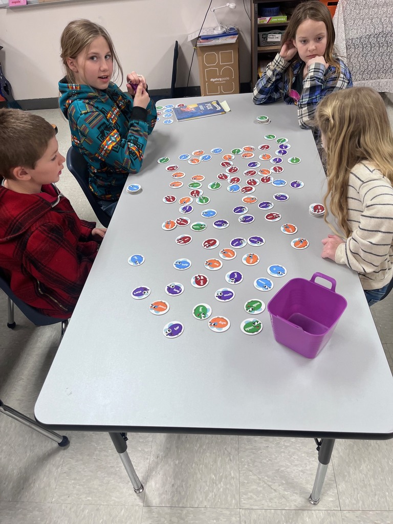 Four children sit around a grey table playing a matching game with dozens of colorful, bug-shaped circular cards spread across the surface.