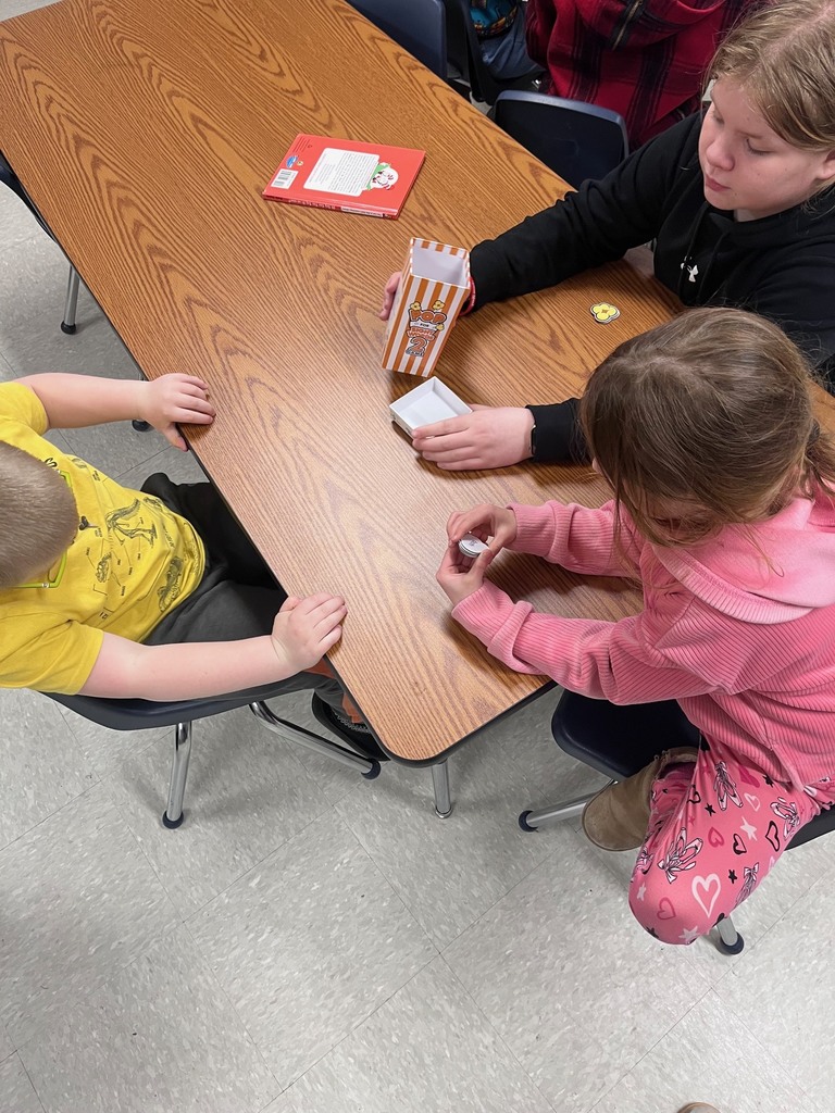 : Three children sit at a wooden desk playing a "Pop for Sight Words" game. One child is reaching into the popcorn-themed box while another examines a small white game piece.