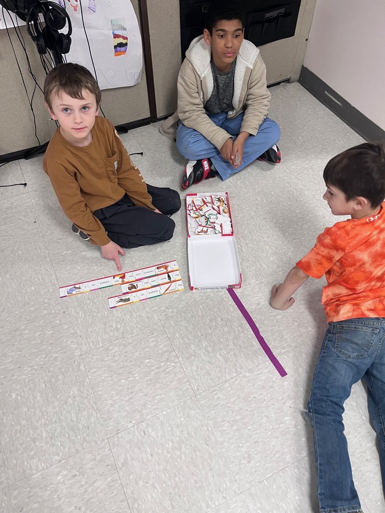 Three boys sit in a circle on the floor working with a "Silly Sentences" game. They are connecting interlocking word pieces to form long, colorful sentence chains. Image 2: A high-angle shot of three girls sitting in a circle on the floor. One girl holds a "Pop for Sight Words" game box while another holds a small circular word tile. Image 3: Three children sit at a wooden desk playing a "Pop for Sight Words" game. One child is reaching into the popcorn-theme