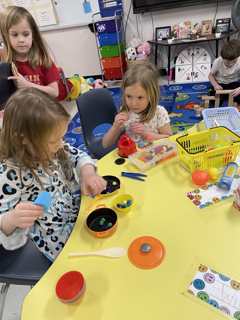 Three children engage in imaginative play at a yellow table. They are using colorful silicone cupcake liners, plastic pots, pans, and small pom-poms to simulate cooking or sorting.