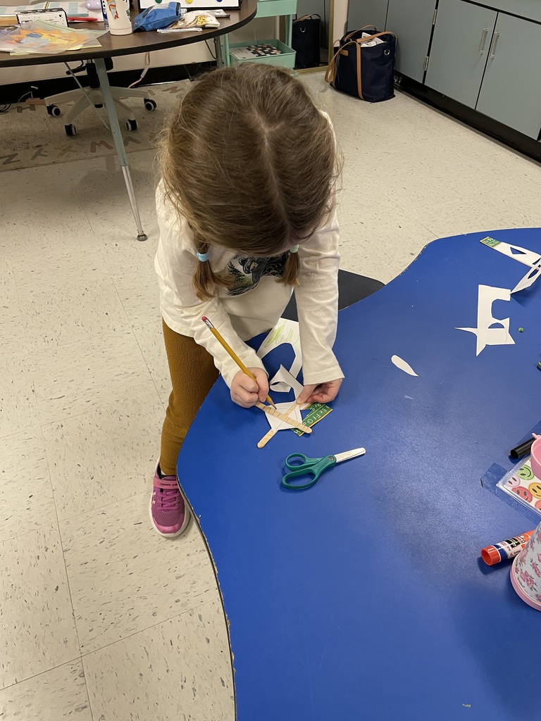 High-angle shot of a young girl with pigtails standing at a blue table. She is using a yellow pencil to carefully trace or mark a white piece of paper that is layered over colorful cards and craft sticks. A pair of green safety scissors lies nearby.