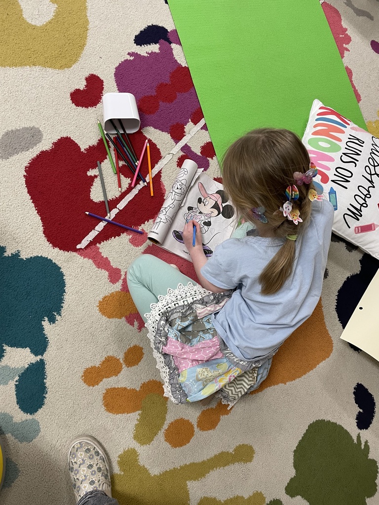 : Overhead view of a girl with colorful butterfly clips in her hair sitting cross-legged on a splatter-patterned rug. She is focused on coloring a picture of Minnie Mouse in a coloring book with a blue colored pencil.