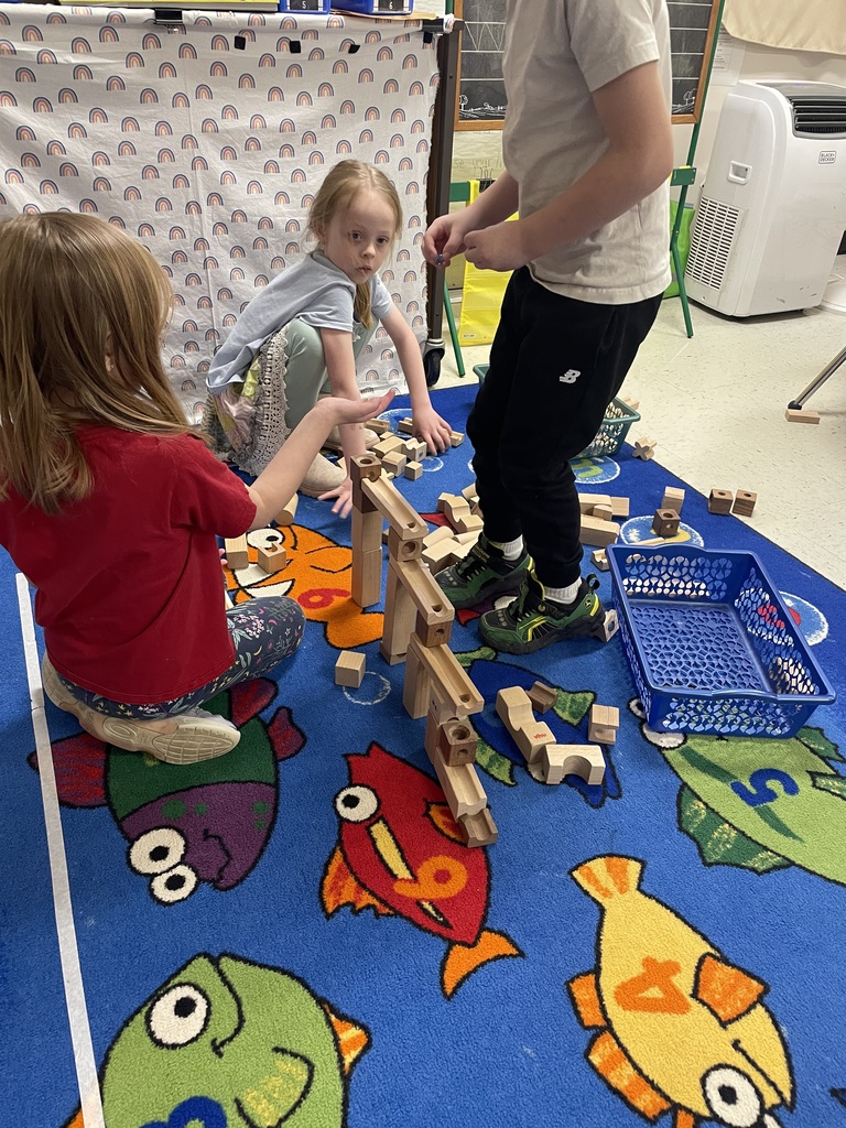 Three children collaborate on a large wooden marble run over a fish-themed rug. One boy stands while two girls sit nearby; they appear to be testing the track with marbles and discussing the construction.