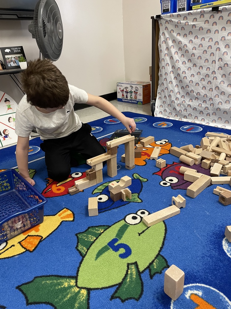 A young boy with brown hair kneels on a blue rug decorated with numbered fish. He is focused on building a tall, intricate marble run using light-colored wooden blocks and tracks.