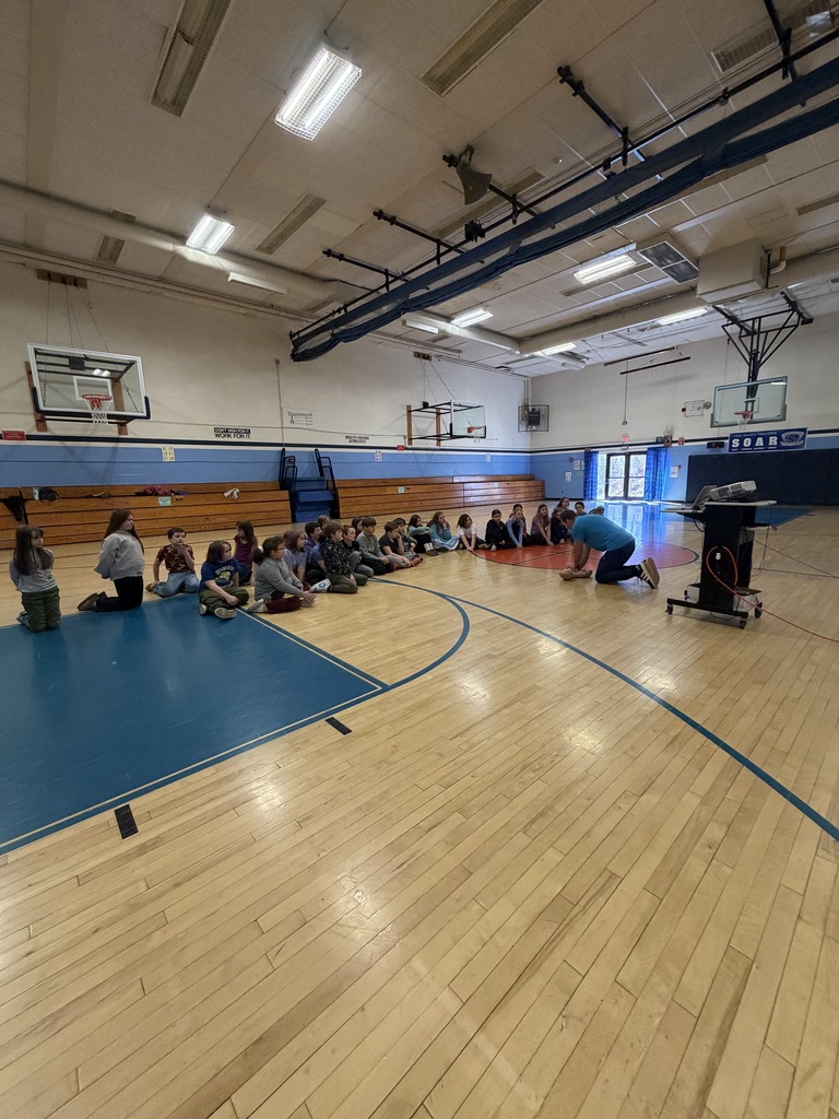Instructor demonstrates CPR technique on a training mannequin as students sit in a semicircle observing in the gym.