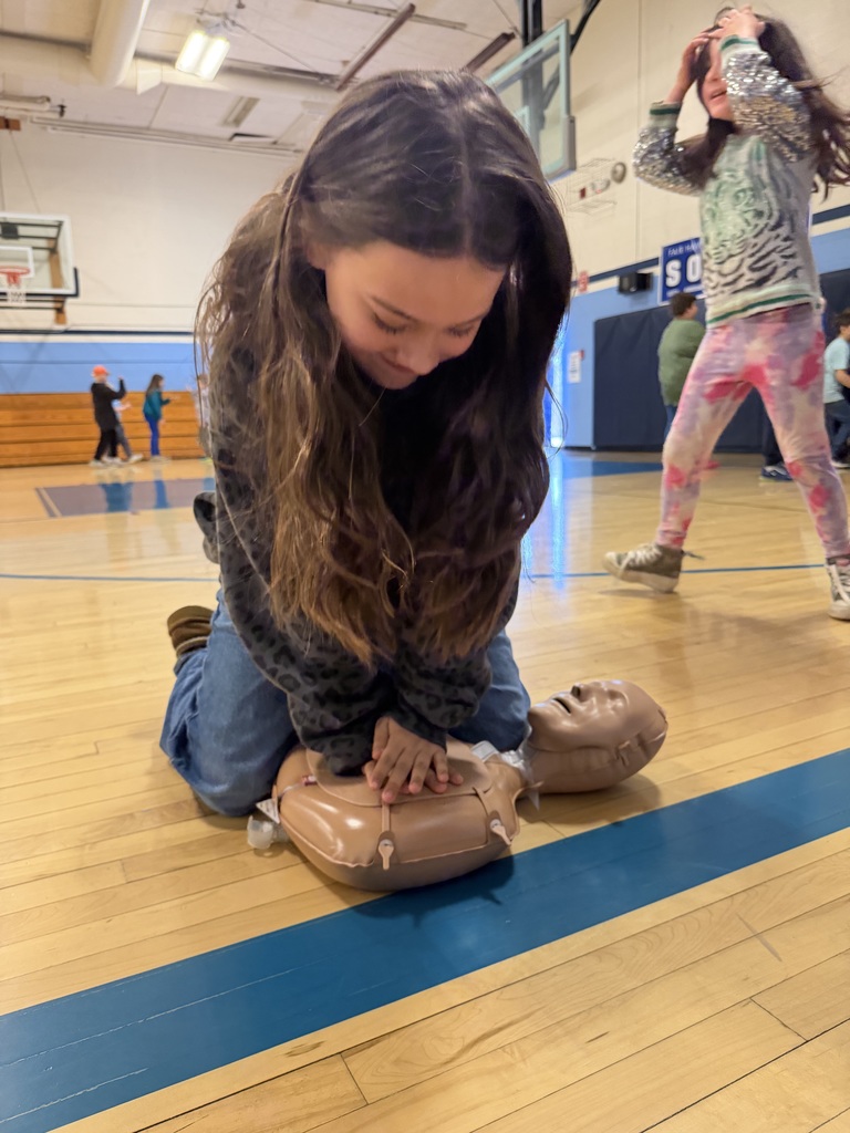 Student kneels on a gym floor practicing chest compressions on a CPR training mannequin while classmates participate in activities nearby.