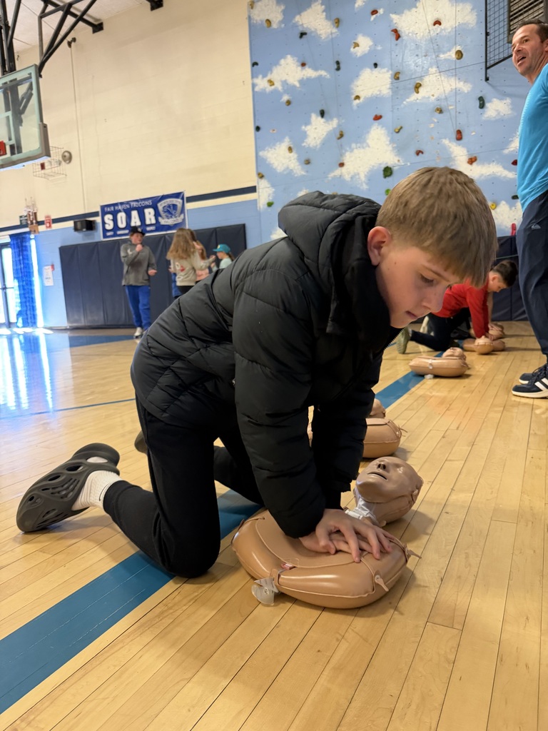 Student performs CPR compressions on a training mannequin while an instructor and classmates practice in the background.