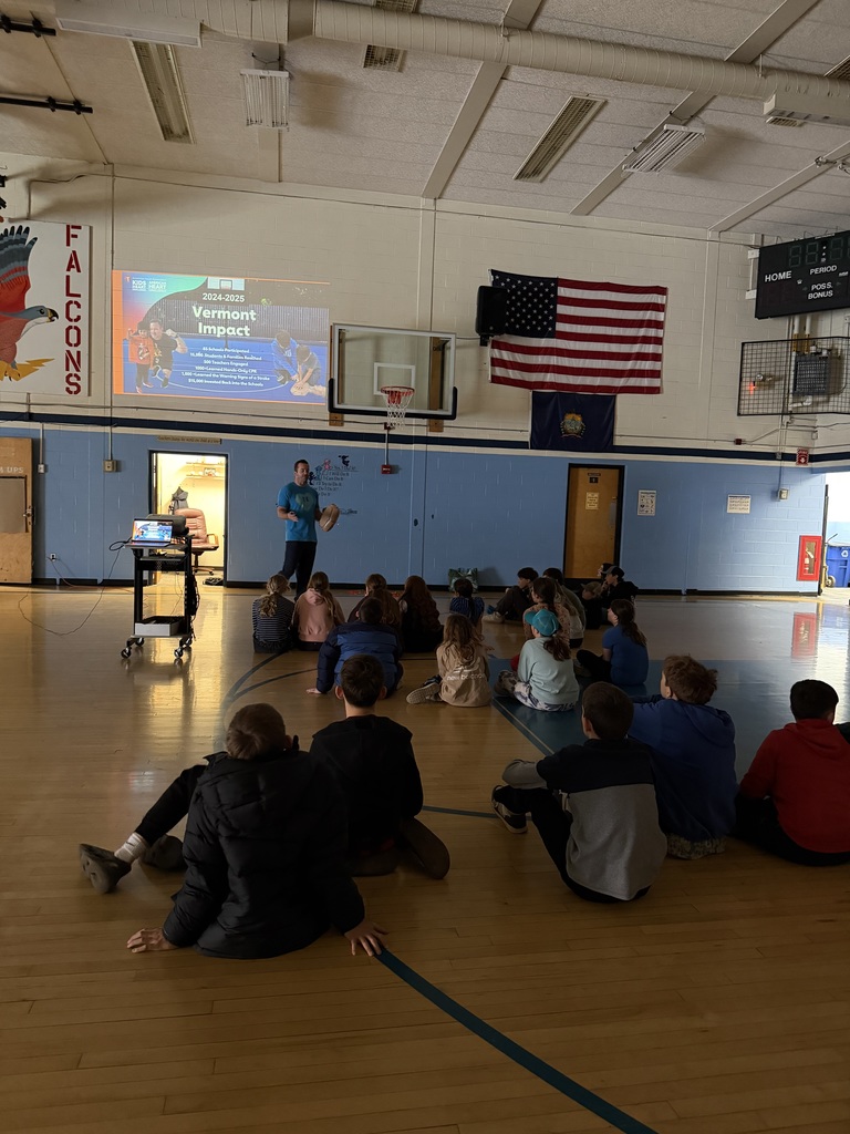 Students sit on the gym floor watching a presenter lead a CPR and emergency response lesson beside a projected screen.