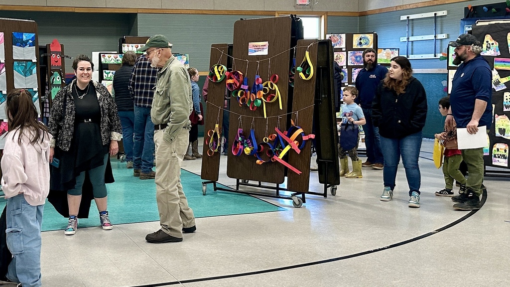 Adults, students, and families walk through a school gym viewing colorful student artwork displayed on panels.