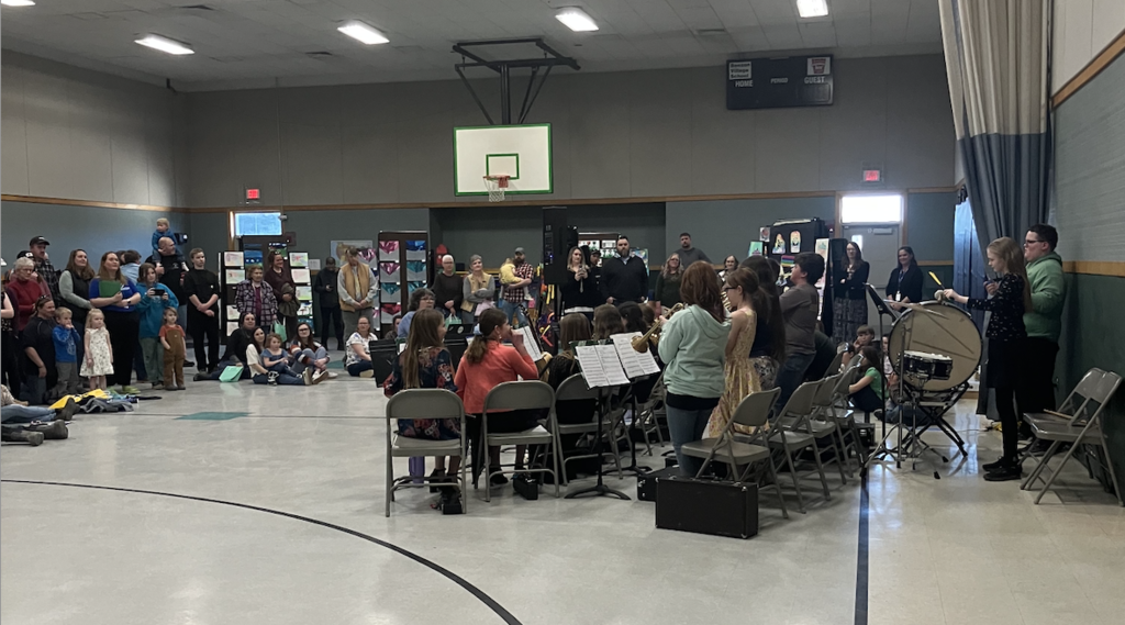 Students perform in a school band seated in a gym while families and community members watch from around the room.